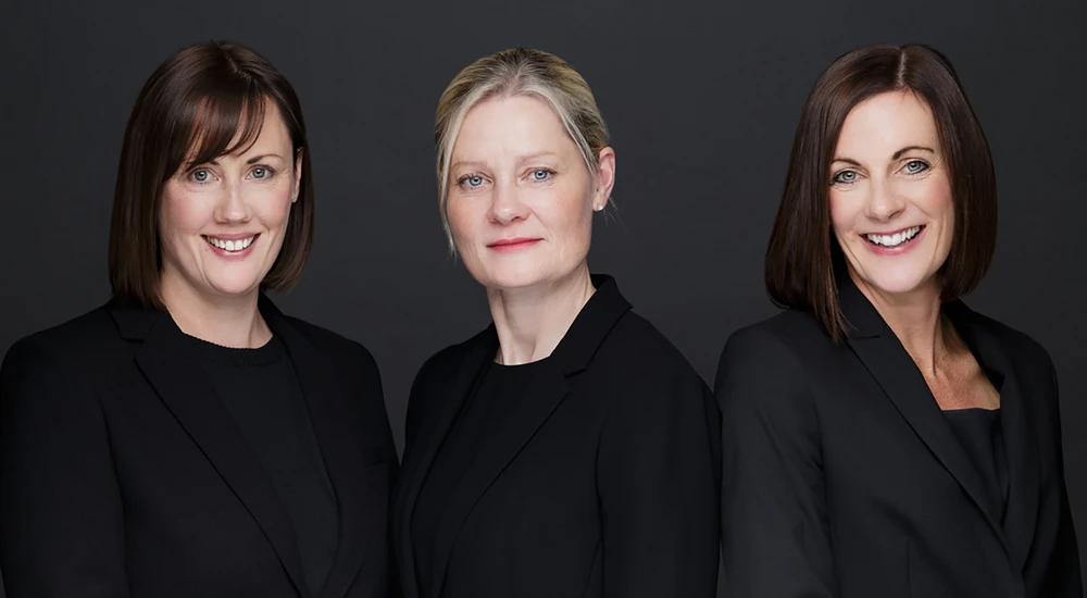 Three professional women in black blazers smiling against a dark background.