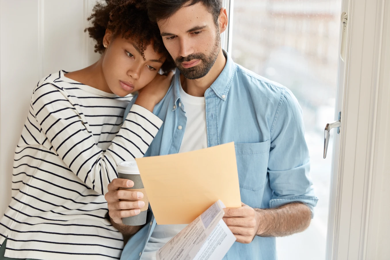 Couple reviewing documents together near a window, with the woman resting her head on the man's shoulder.