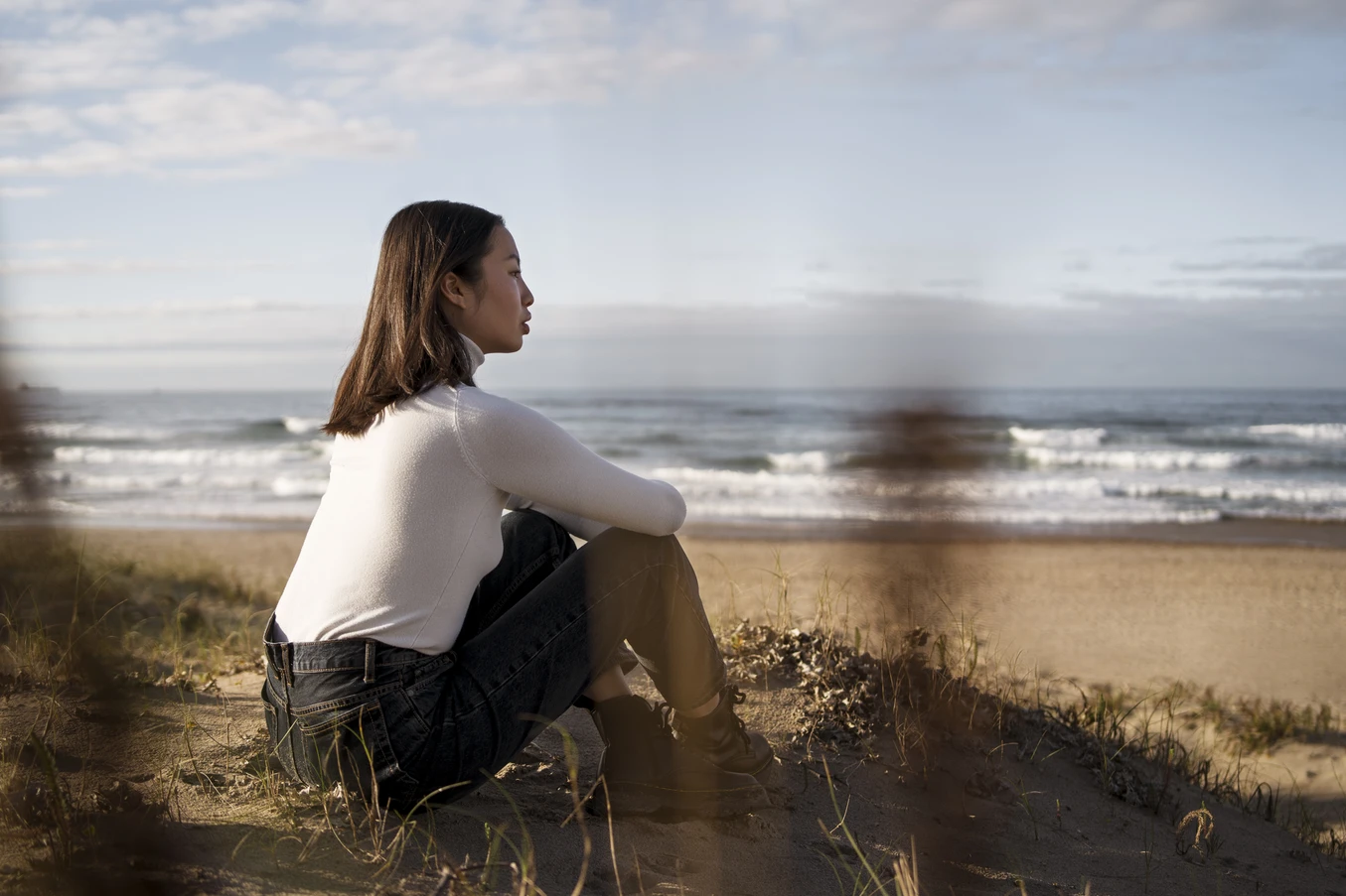 Woman in white sweater and black jeans sitting on sand dunes looking at the ocean under a partly cloudy sky.