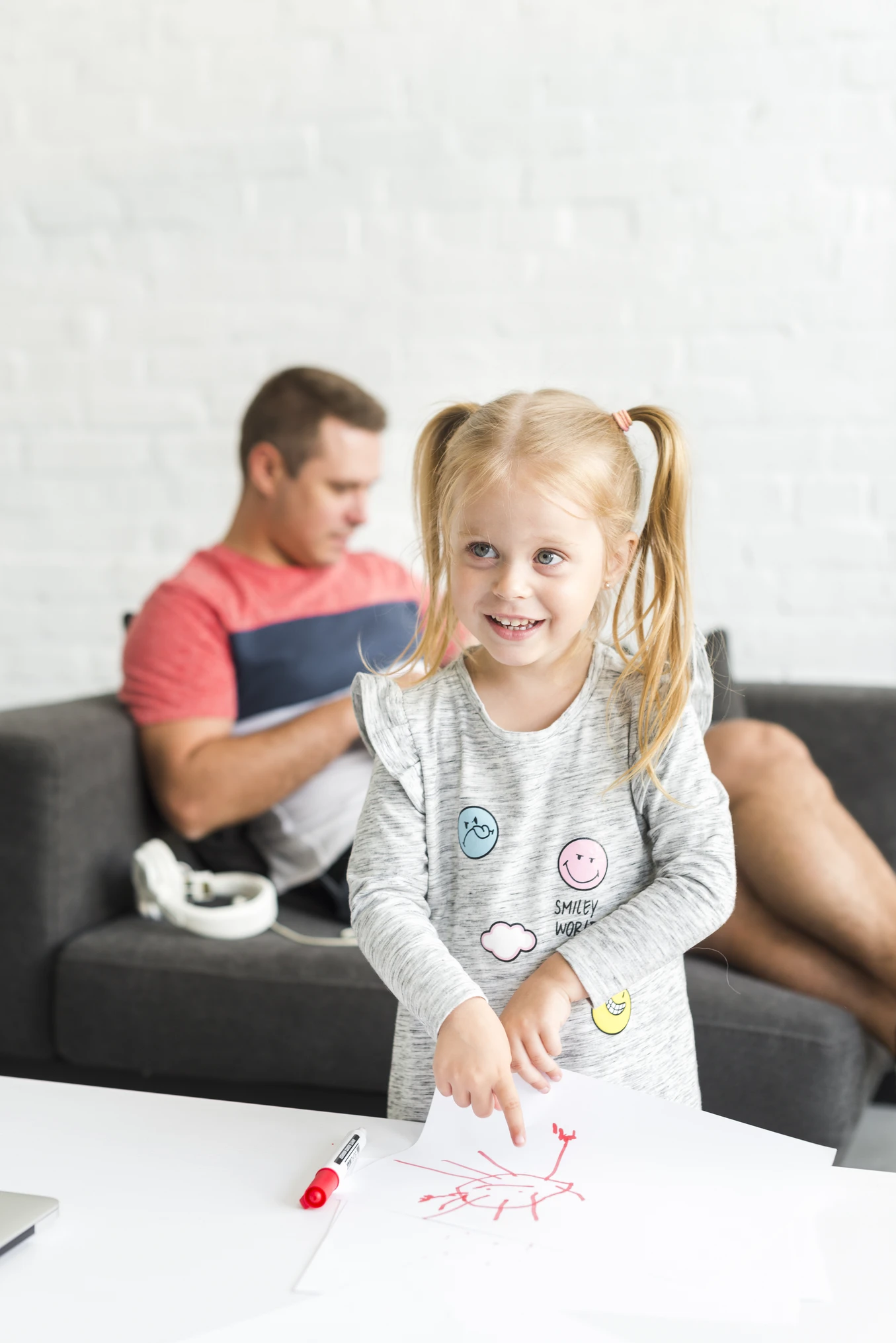 Young girl with blonde pigtails smiling and pointing at a red drawing on white paper, with a man in a red and navy shirt sitting on a couch in the background.