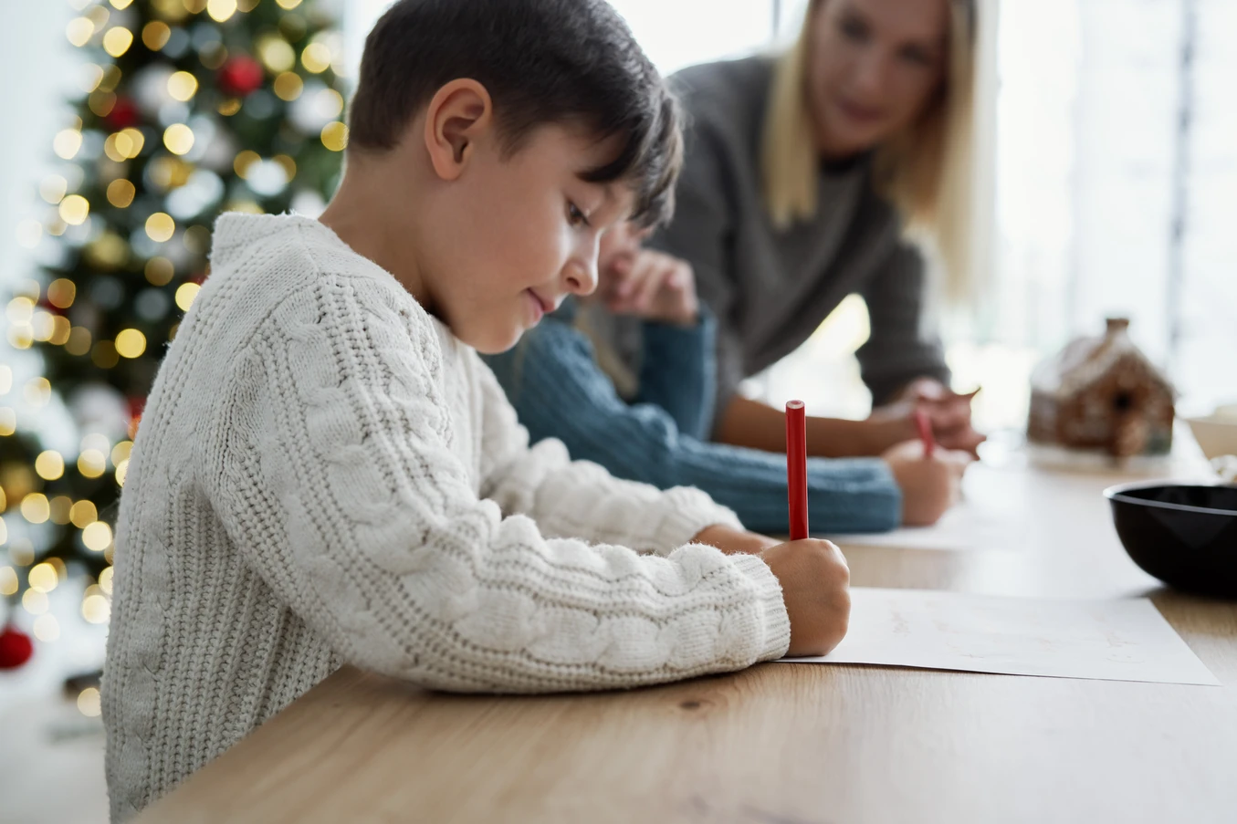 Young boy in a white cable-knit sweater writing with a red pen at a table, with a Christmas tree and a woman blurred in the background.