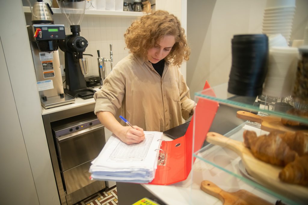 A person with curly hair writing in a notebook while standing behind a counter in a cafe, with a coffee machine and pastries in the background.