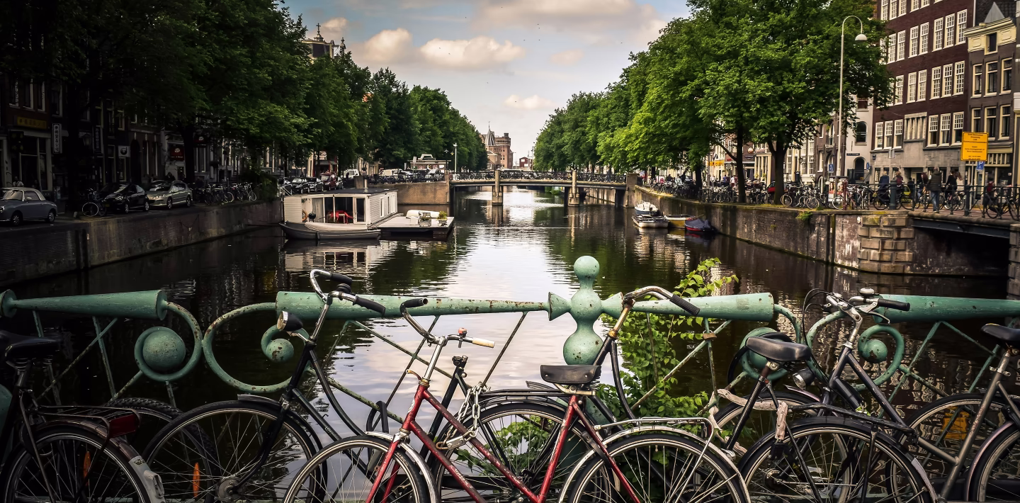 Fietsen geparkeerd bij een groene brug over een gracht met boten en bomen in Amsterdam.