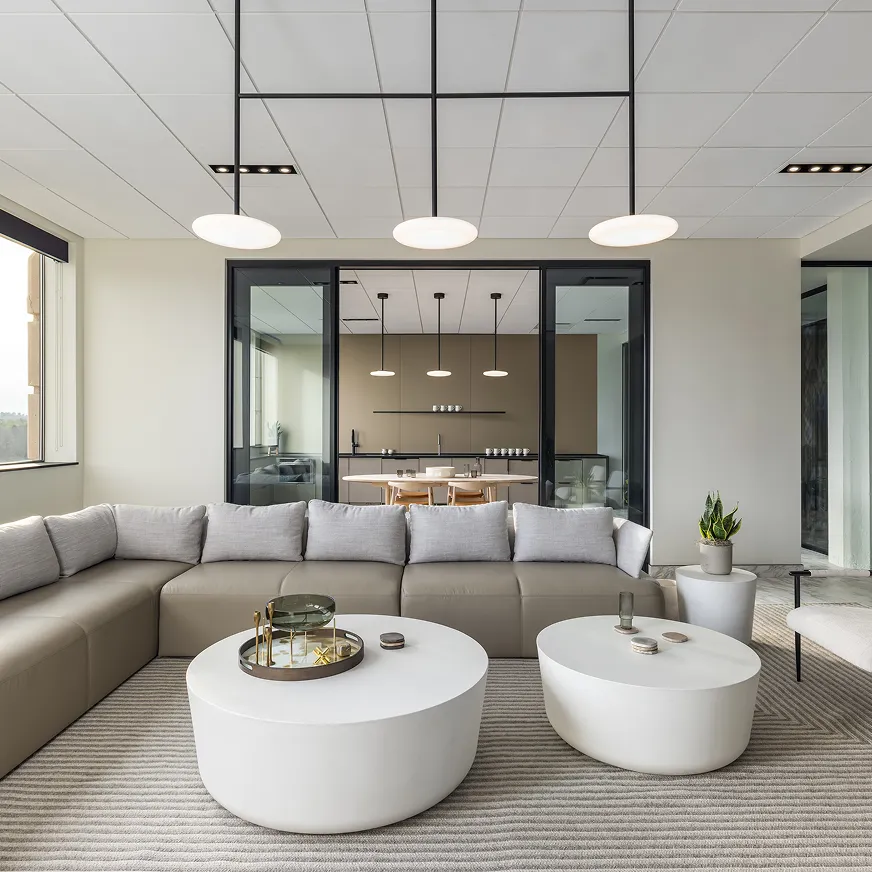 Office lounge area featuring a gray sectional sofa with round white coffee tables and modern lighting.