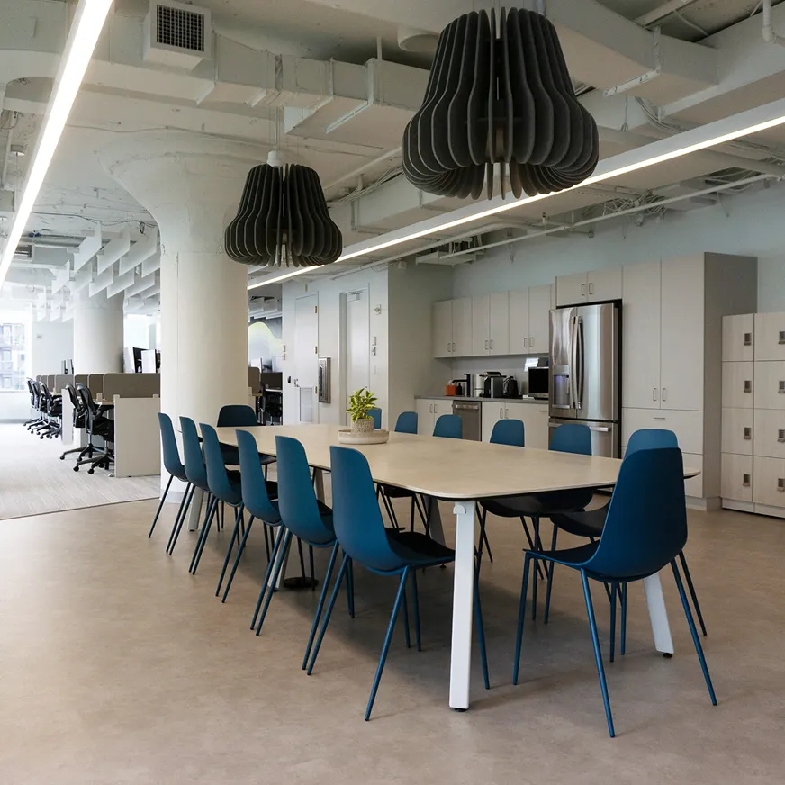 Modern office breakout space with a long table and blue chairs under contemporary ceiling lighting.