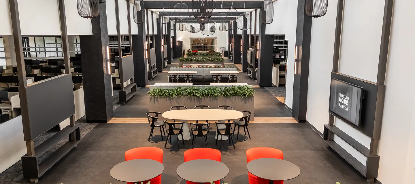 Spacious modern office interior with collaborative dining tables, red chairs, greenery, and open lounge areas for employees.