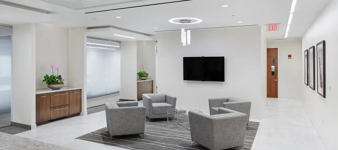 Modern office lobby with four gray lounge chairs around a small table, TV mounted on the wall, and wooden cabinets with plants.