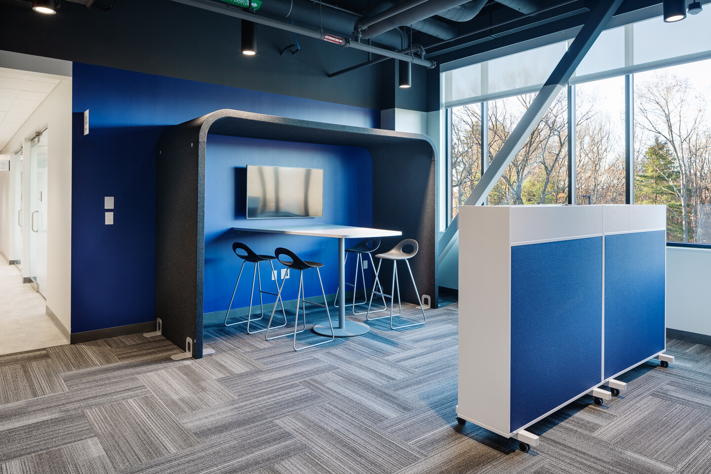 Modern blue office lounge with a built-in meeting nook, bar-height table, stools, and large windows.
