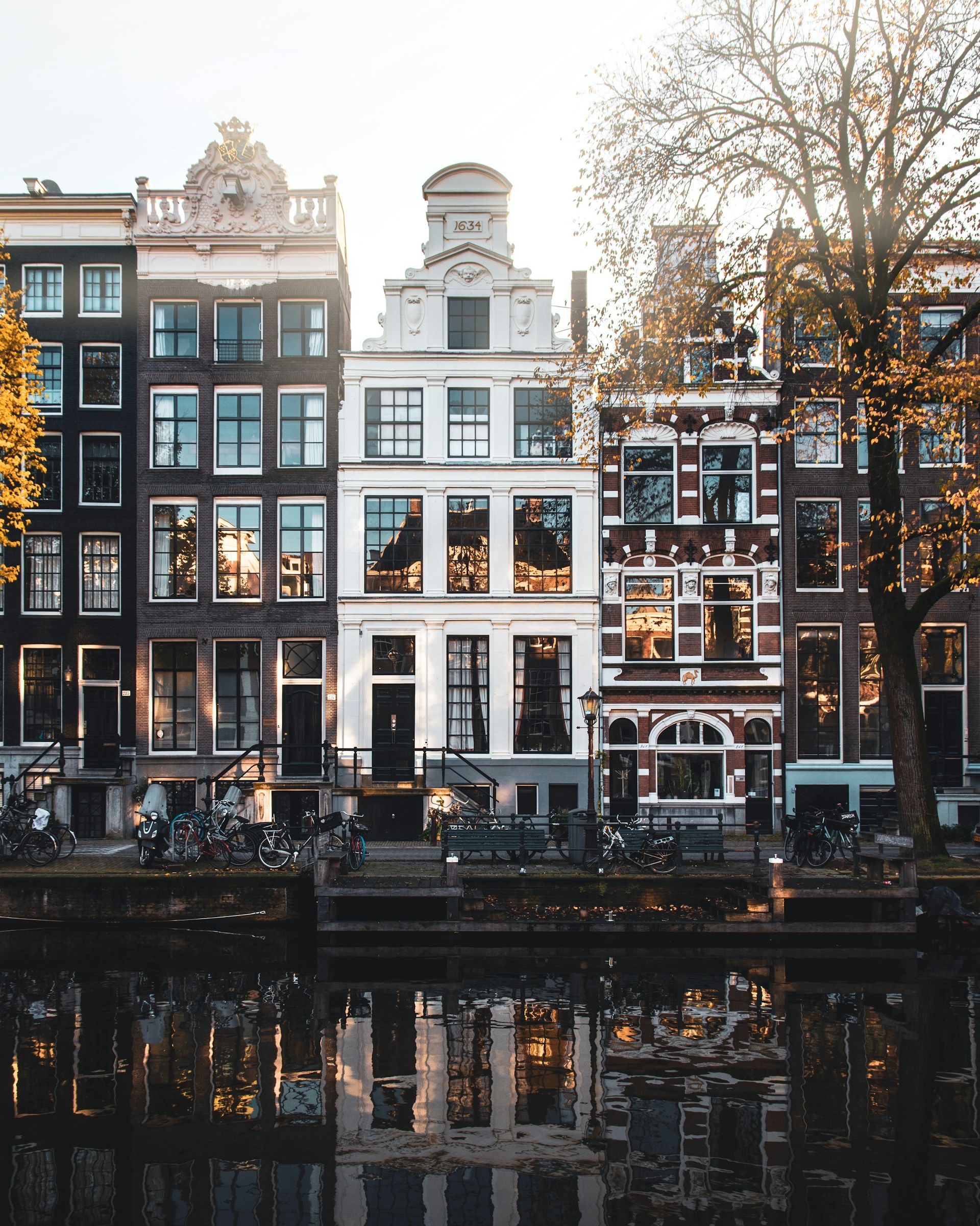 Canal-side traditional Amsterdam buildings with large windows and bicycles parked along the street, reflected in the water.