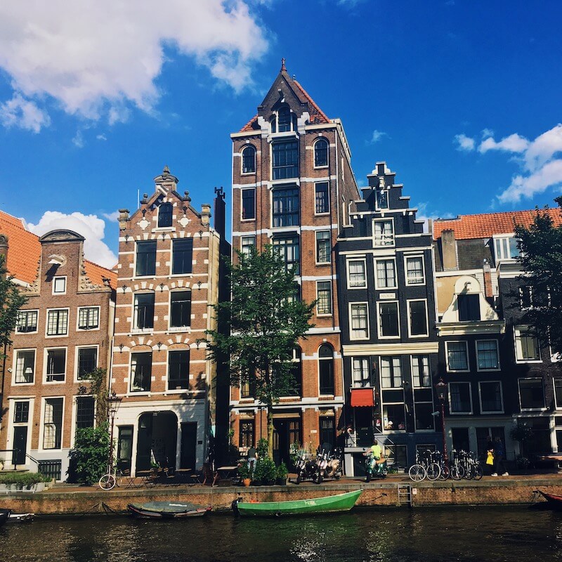 Traditional narrow Amsterdam canal houses with gabled roofs along a waterway under a blue sky.