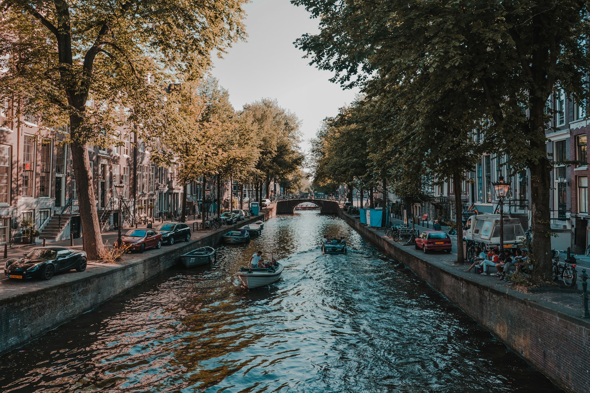 View of an Amsterdam canal with boats, lined by trees and traditional buildings, under a clear sky.