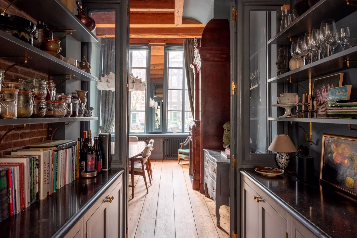 Narrow kitchen passage with shelves holding jars, books, and glassware leading to a dining area with wooden floor and large windows.