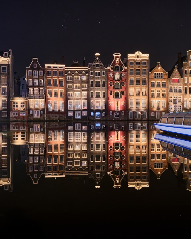 Tall, narrow Amsterdam canal houses illuminated at night with their reflections in calm water and a docked boat on the right.