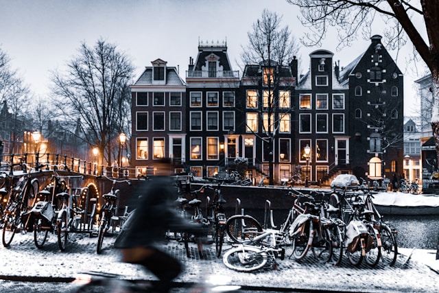 Snow-covered canal in Amsterdam with bicycles parked along the railing and a blurred cyclist in motion in the foreground during twilight.