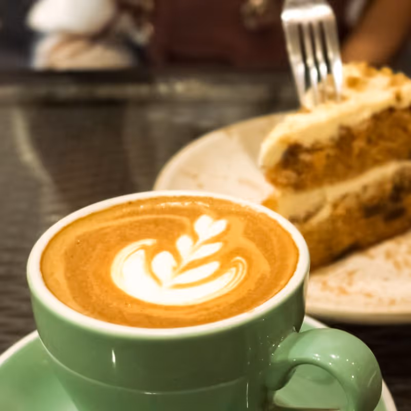 Latte mit Blattmuster in grüner Tasse neben einem Stück Kuchen