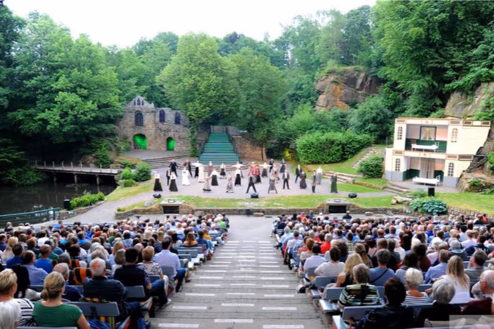 Freilichtbühne mit Publikum und Schauspielern vor historischer Kulisse im Grünen