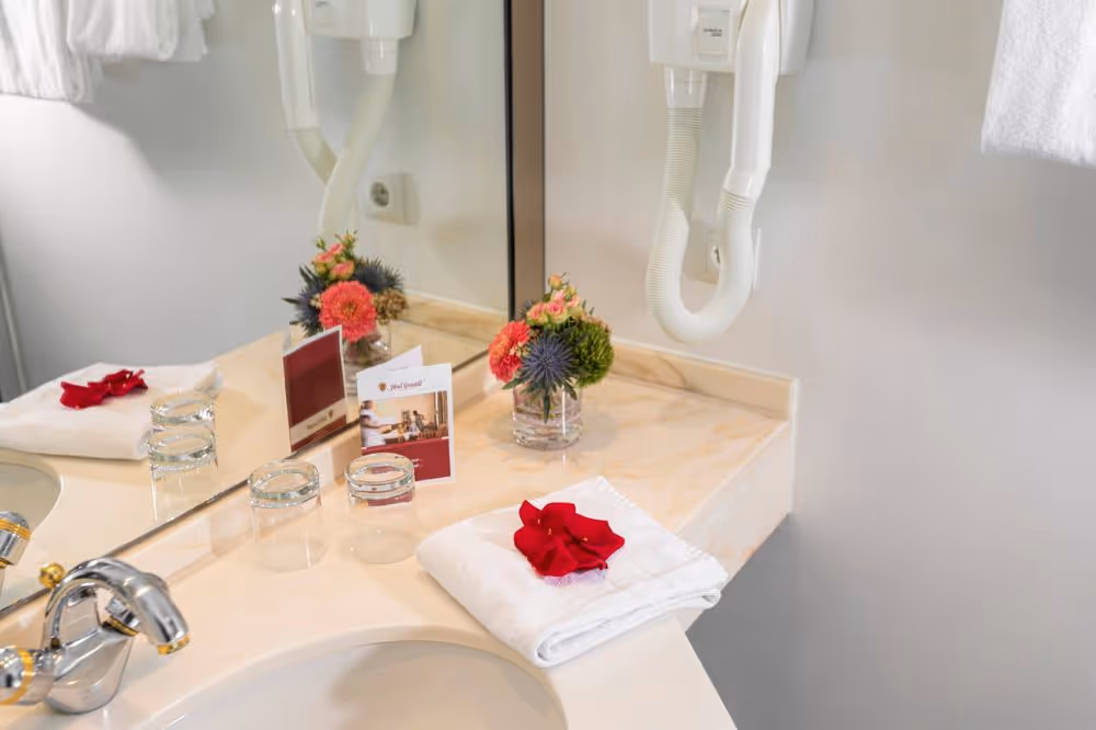 Badezimmerwaschbecken mit Blumen, Gläsern und roter Blüte auf Handtuch