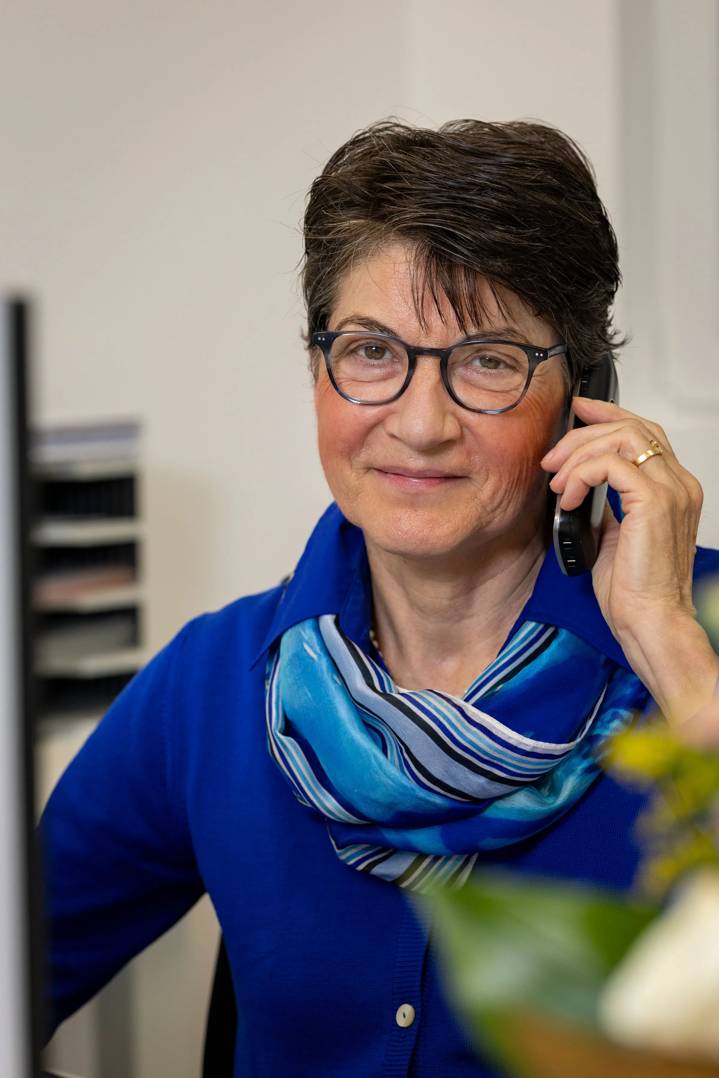 Ältere Frau mit Brille und blauem Schal, die am Telefon spricht und freundlich lächelt.
