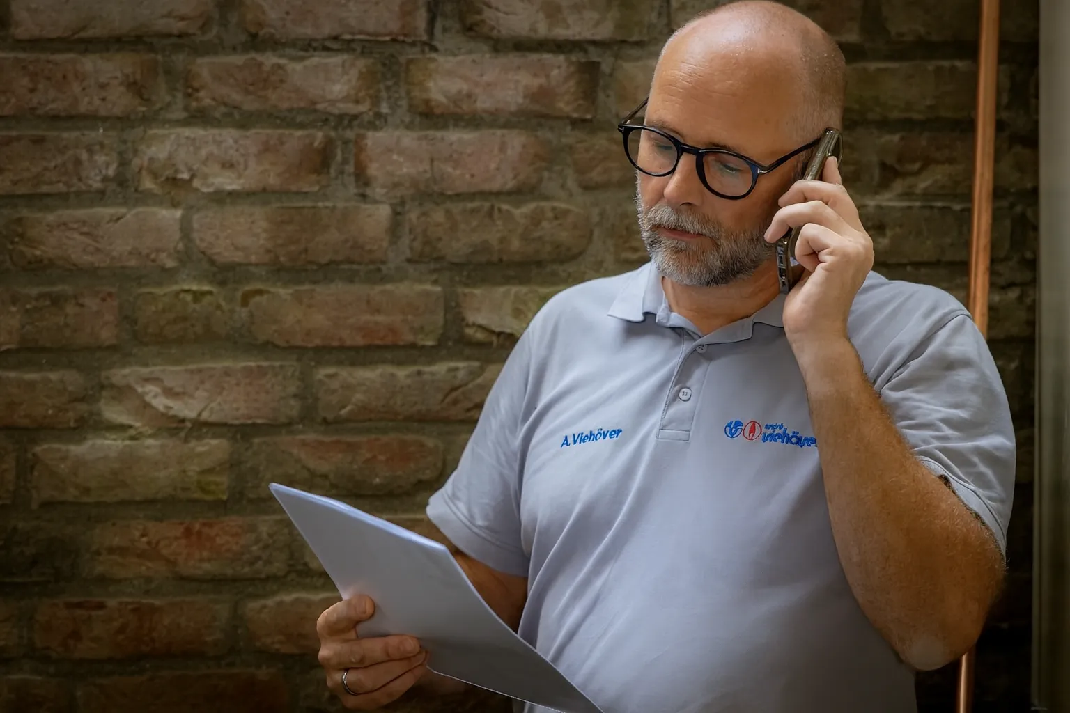 Mann mit Brille und grauem Bart spricht am Telefon und liest ein Dokument vor einer Backsteinwand.
