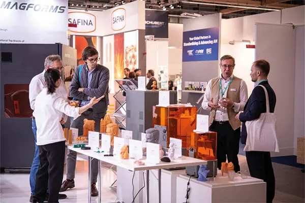 Menschen unterhalten sich an einem Messestand mit orangefarbenen 3D-Druckern und gedruckten Modellen auf Tischen.