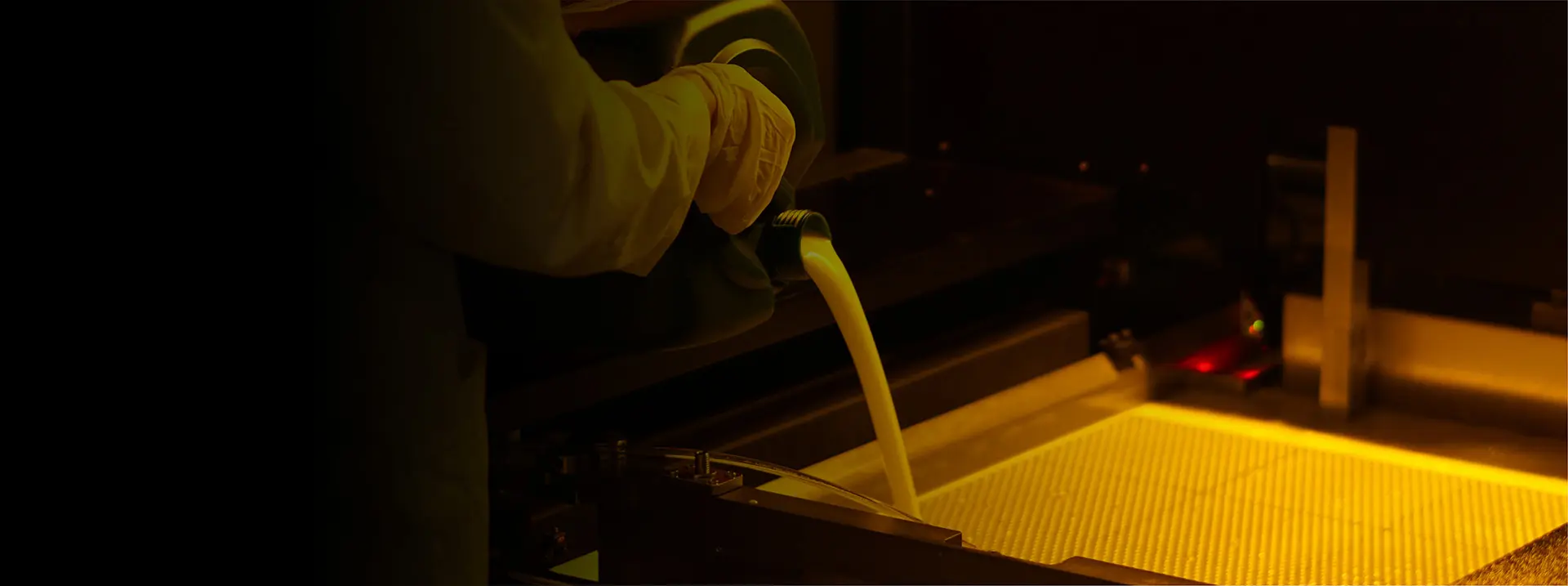 Person in Schutzkleidung gießt gelbliche Flüssigkeit in eine Laborausrüstung unter gelbem Licht.