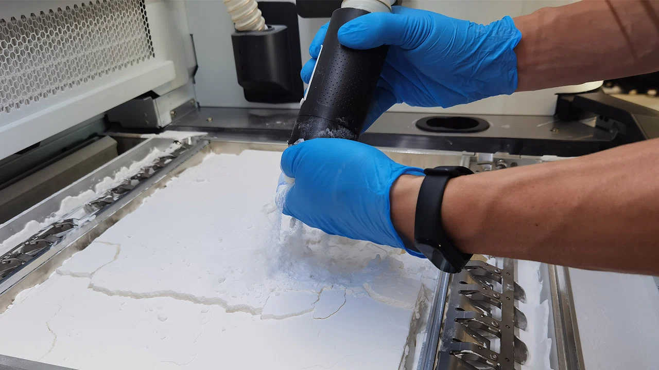 Person mit blauen Handschuhen, der Pulver in eine Maschine streut, vermutlich für die additive Fertigung.