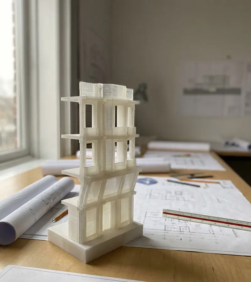 3D-gedrucktes Architekturmodell eines mehrstöckigen Gebäudes auf einem Tisch mit Bauplänen und Zeichengeräten.