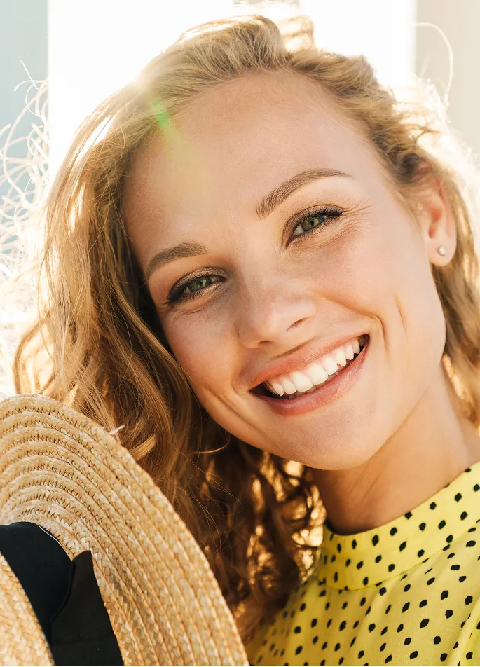 Close-up of a smiling woman with curly blonde hair holding a straw hat, wearing a yellow polka dot top, with sunlit background.