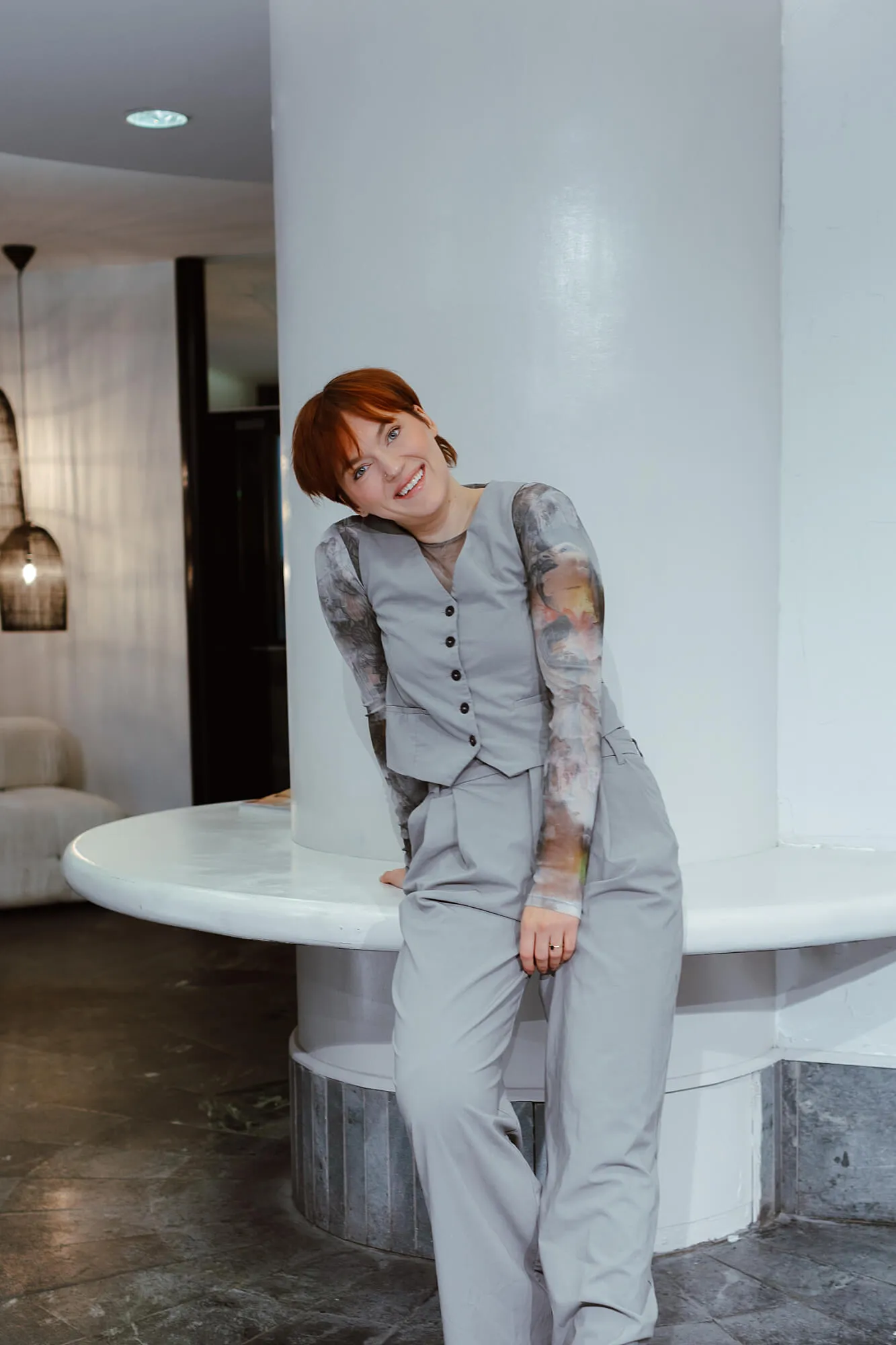 Smiling woman with short red hair wearing a gray vest and printed long-sleeve shirt leaning against a white circular bench inside a modern room.