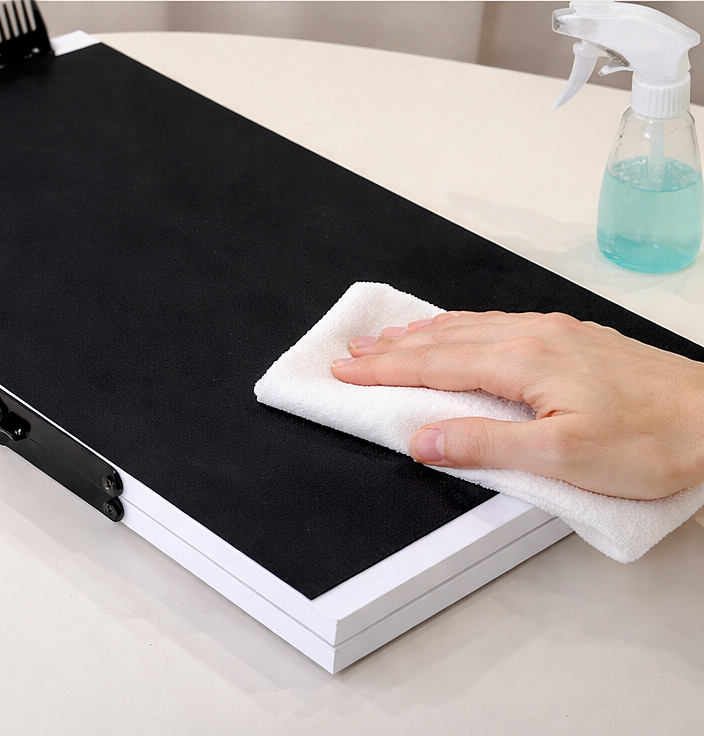 Hand wiping a black clipboard with a white cloth on a white surface, next to a spray bottle with blue liquid.