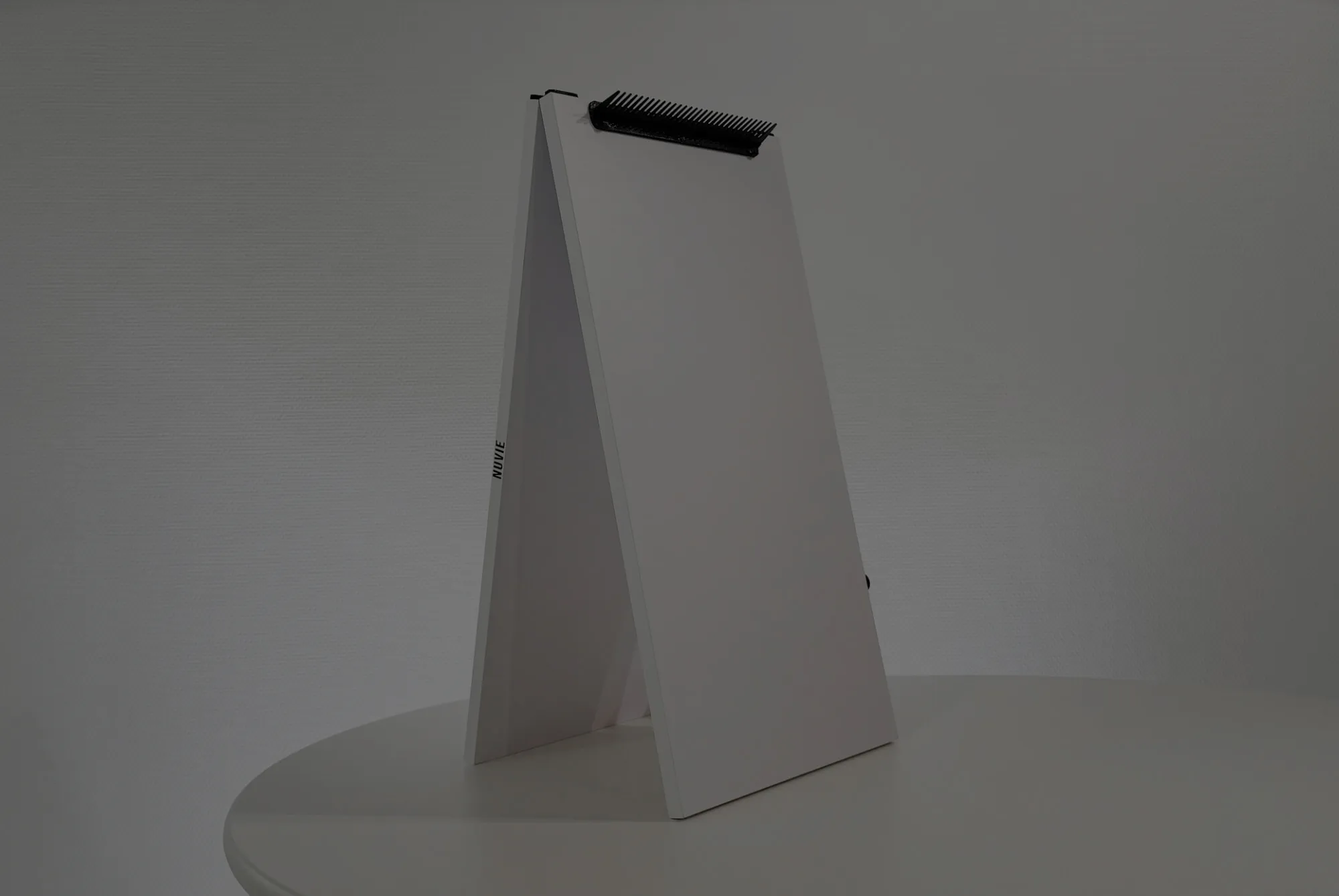 A white standing memo pad with a black comb attached at the top, positioned on a round white table against a plain gray background.