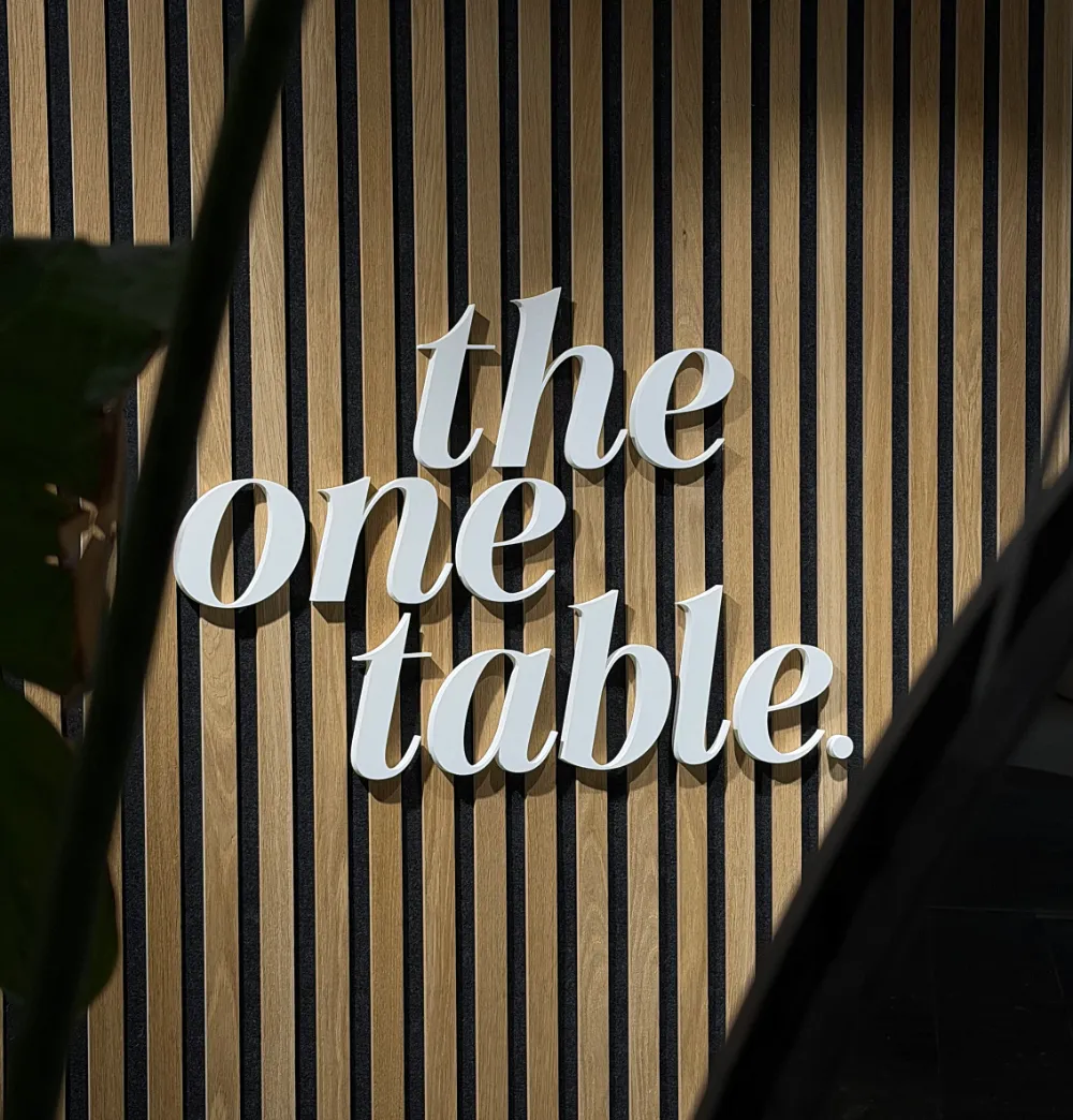 White 3D letters on a wooden slat wall spelling 'the one table.'