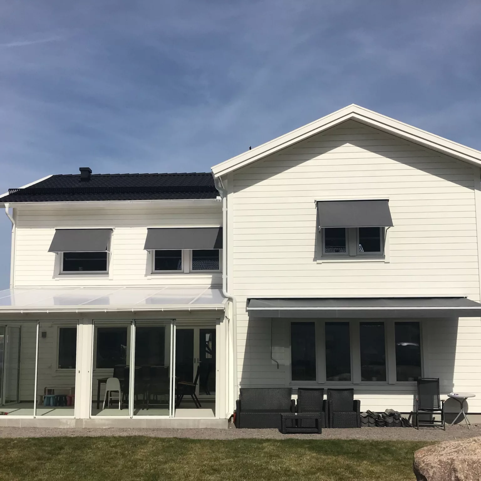 Vit tvåvåningsvilla med grå fönstermarkiser F320 och en terrassmarkis T220, glasat uterum och utemöbler på gräsmatta under blå himmel i Stockholm.