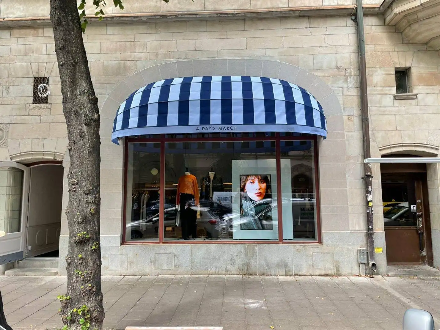 Skyltfönster med randig blå och vit korgmarkis K200 och en skylt med texten A DAY'S MARCH ovanför, visar kläder och ett porträttfoto av en kvinna i Stockholm.