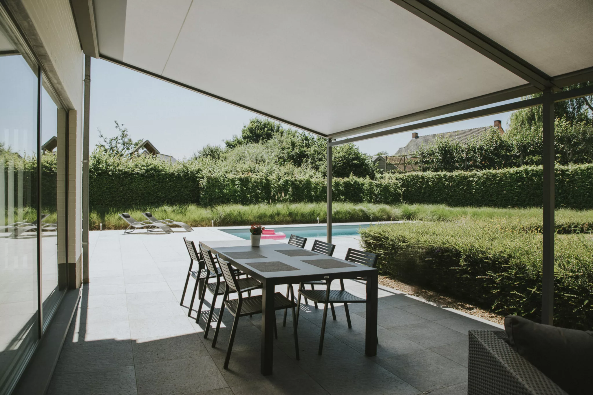 Uteplats med matbord och sex stolar under en pergola vid en pool omgiven av häckar och grönska i Stockholm.