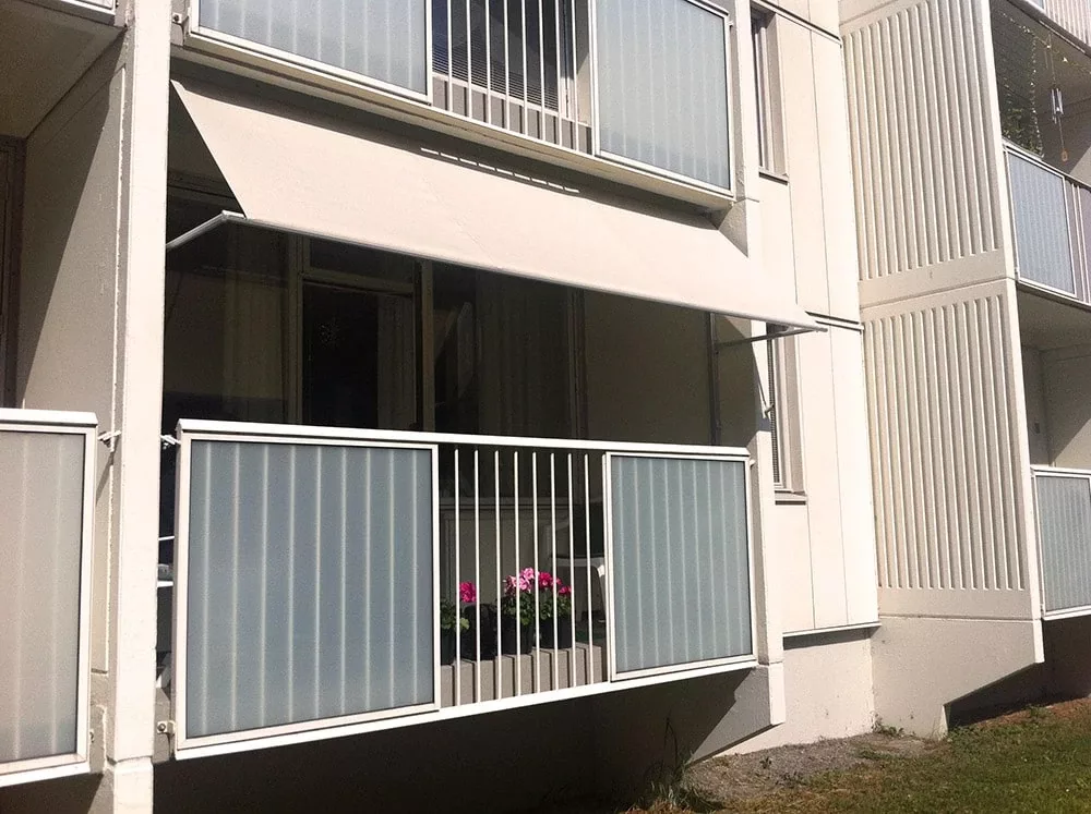 Ljus balkong med frostat glasräcke, beige balkongmarkis B260 och rosa blommor i blomlåda på en vit byggnad i Stockholm.