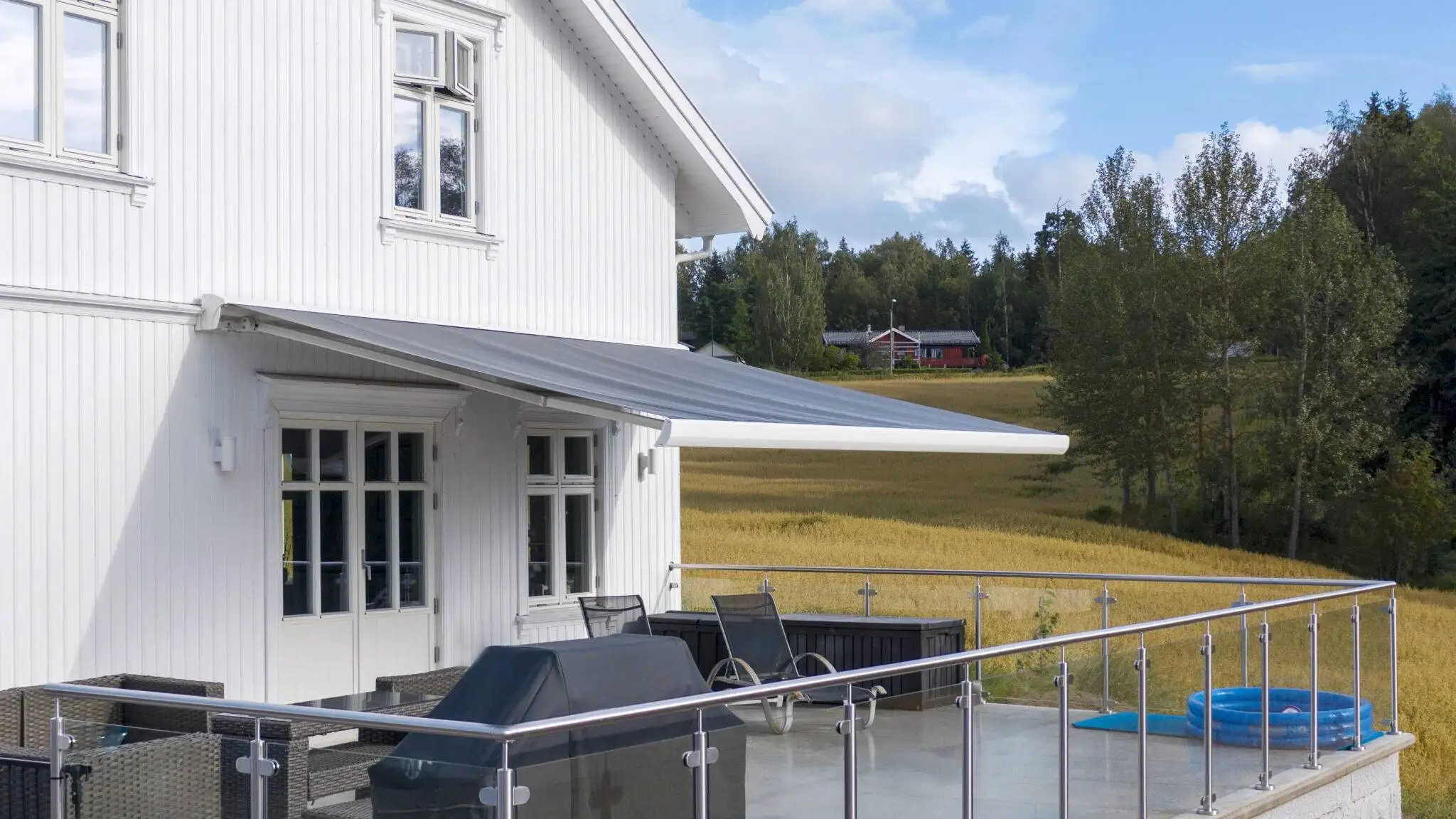 Uteplats med glasräcke vid ett vitt trähus, med en utfälld terrassmarkis ovanför dörren och träd i bakgrunden i Stockholm.