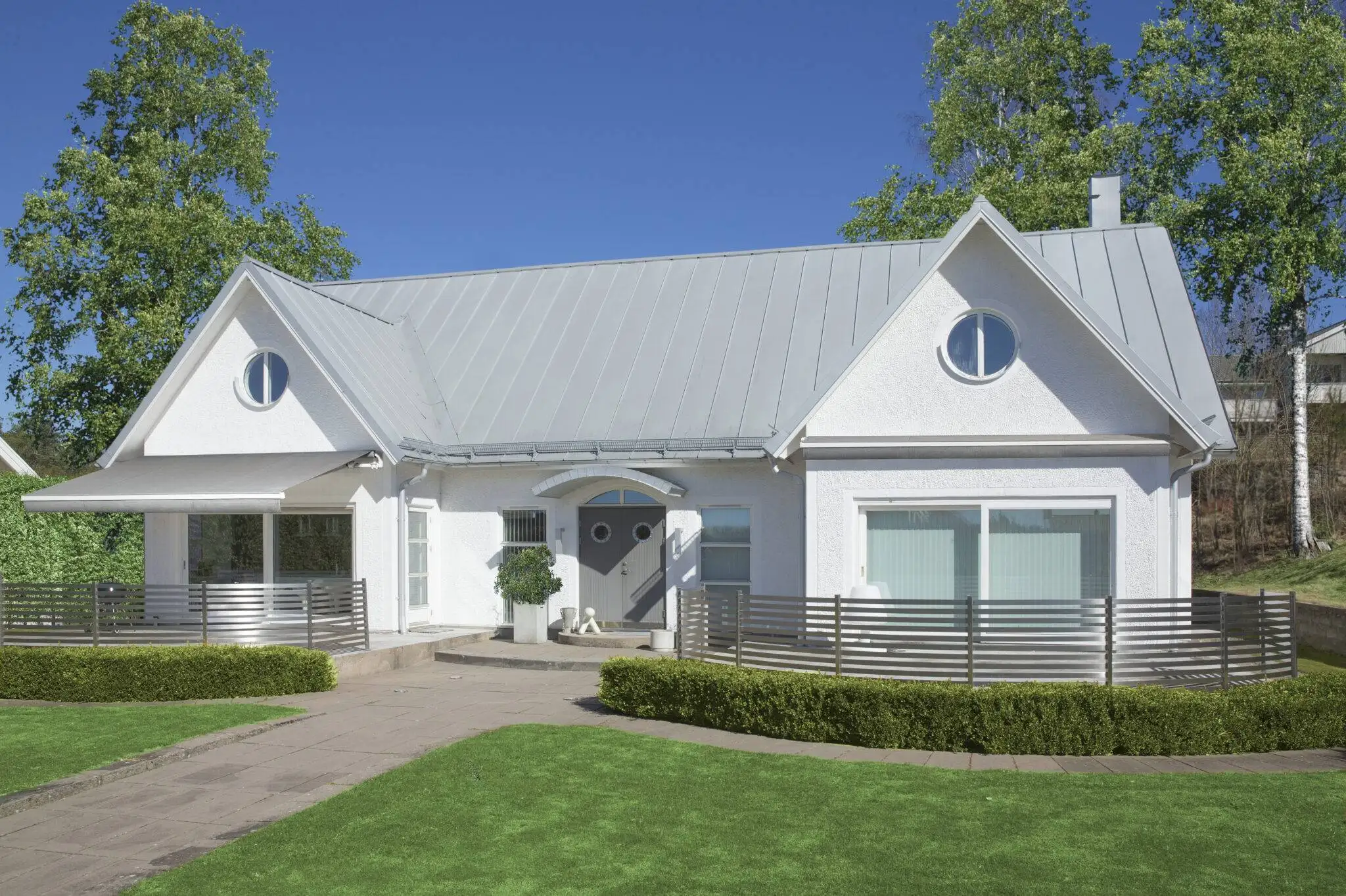 Vit enplansvilla med grått plåttak, rundade fönster vid taknockarna, terrass med metallräcke och välskött gräsmatta och buskar.