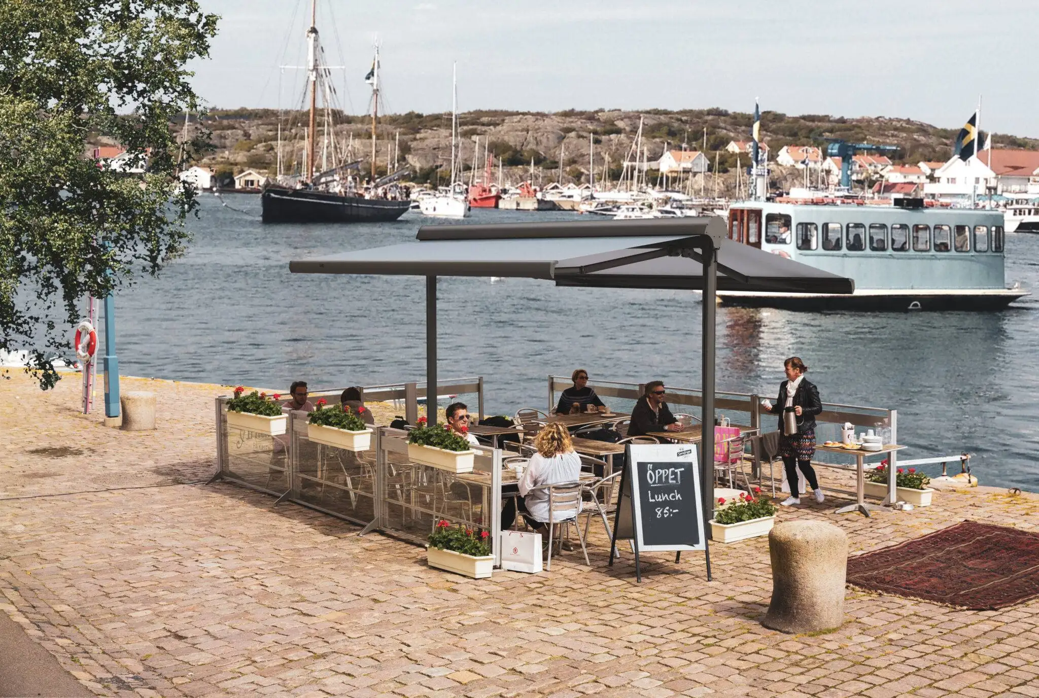 Uteservering vid vattnet med personer som sitter under en terrassmarkis Duo, bakom ligger segelbåtar och en passagerarbåt i Stockholm.