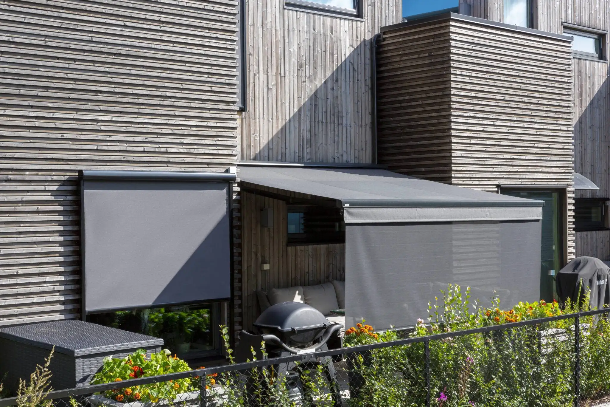 En modern träfasad med en nedrullad zipscreen Z460, en utfälld terrassmarkis T420, en grill och en uteplats med soffa bakom markiserna i Stockholm.