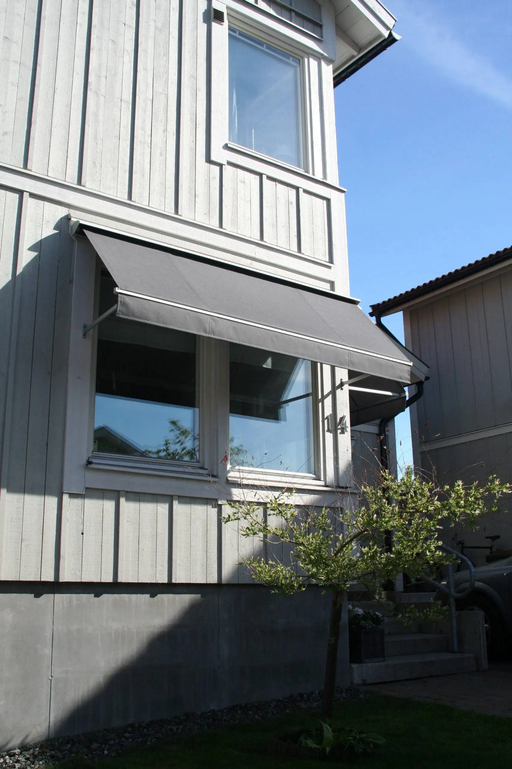 Grå fönstermarkis F320 ovanför två fönster på en vit träfasad med en liten buske framför huset i Stockholm.