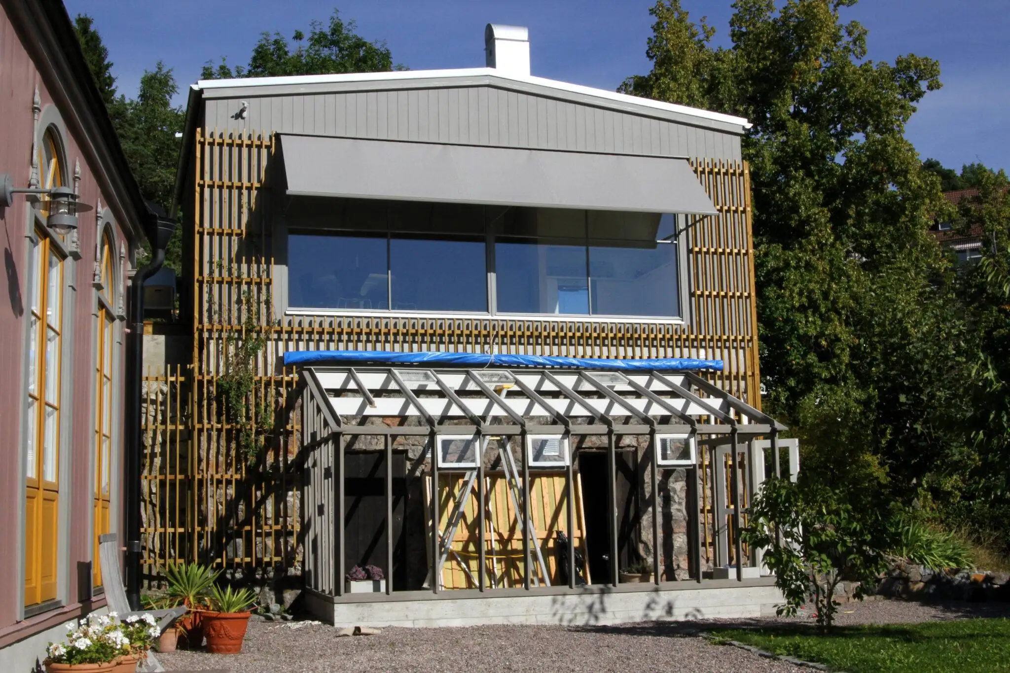 Ett pågående byggprojekt av ett växthus med öppen ramställning framför ett tvåvåningshus med stora fönster och grå fönstermarkis F420 i Stockholm.