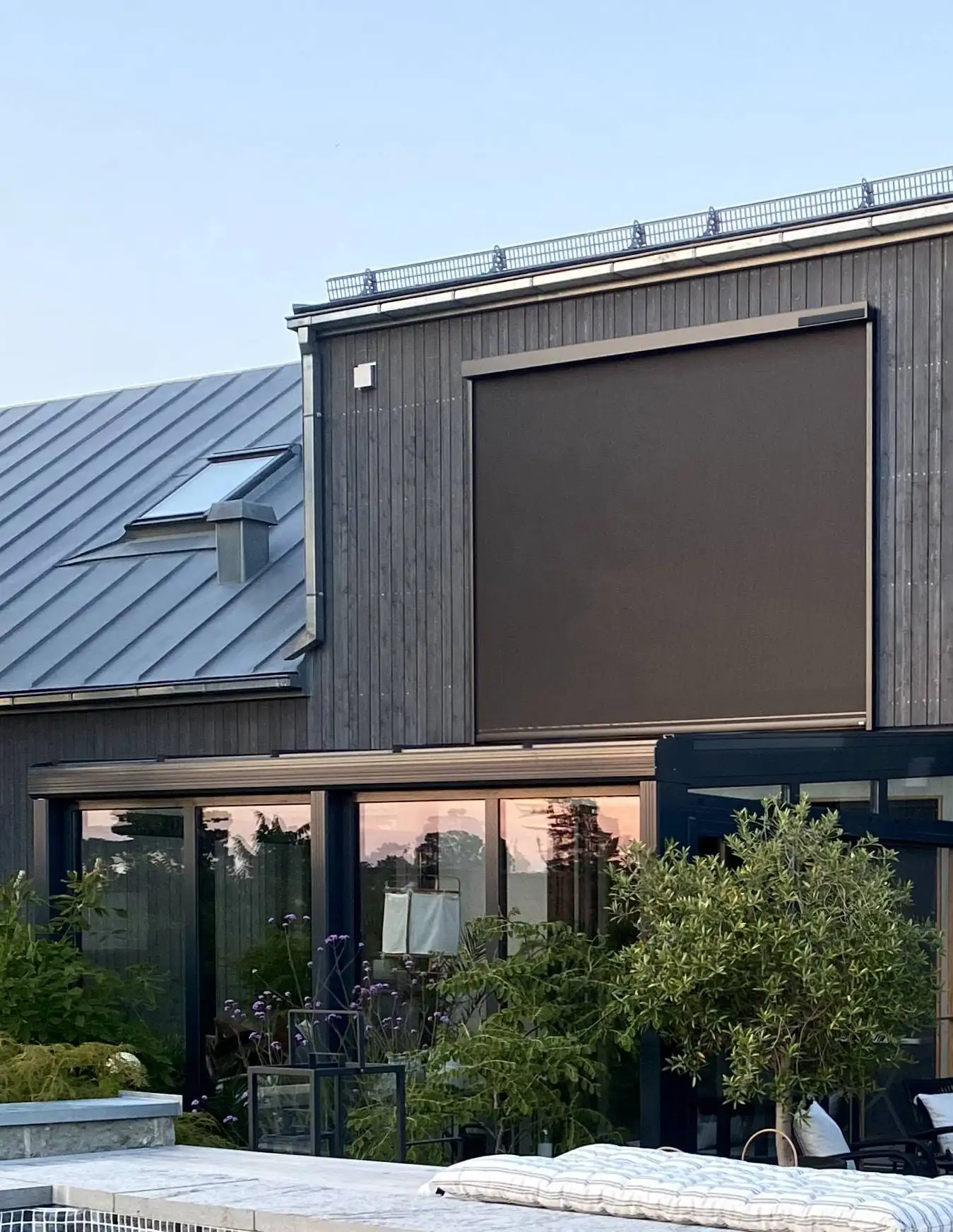 Modernt hus med mörk träfasad, stora fönster och en nedrullad mörk zipscreen Z460 4K på övervåningen, med en pool och växter i förgrunden i Stockholm.