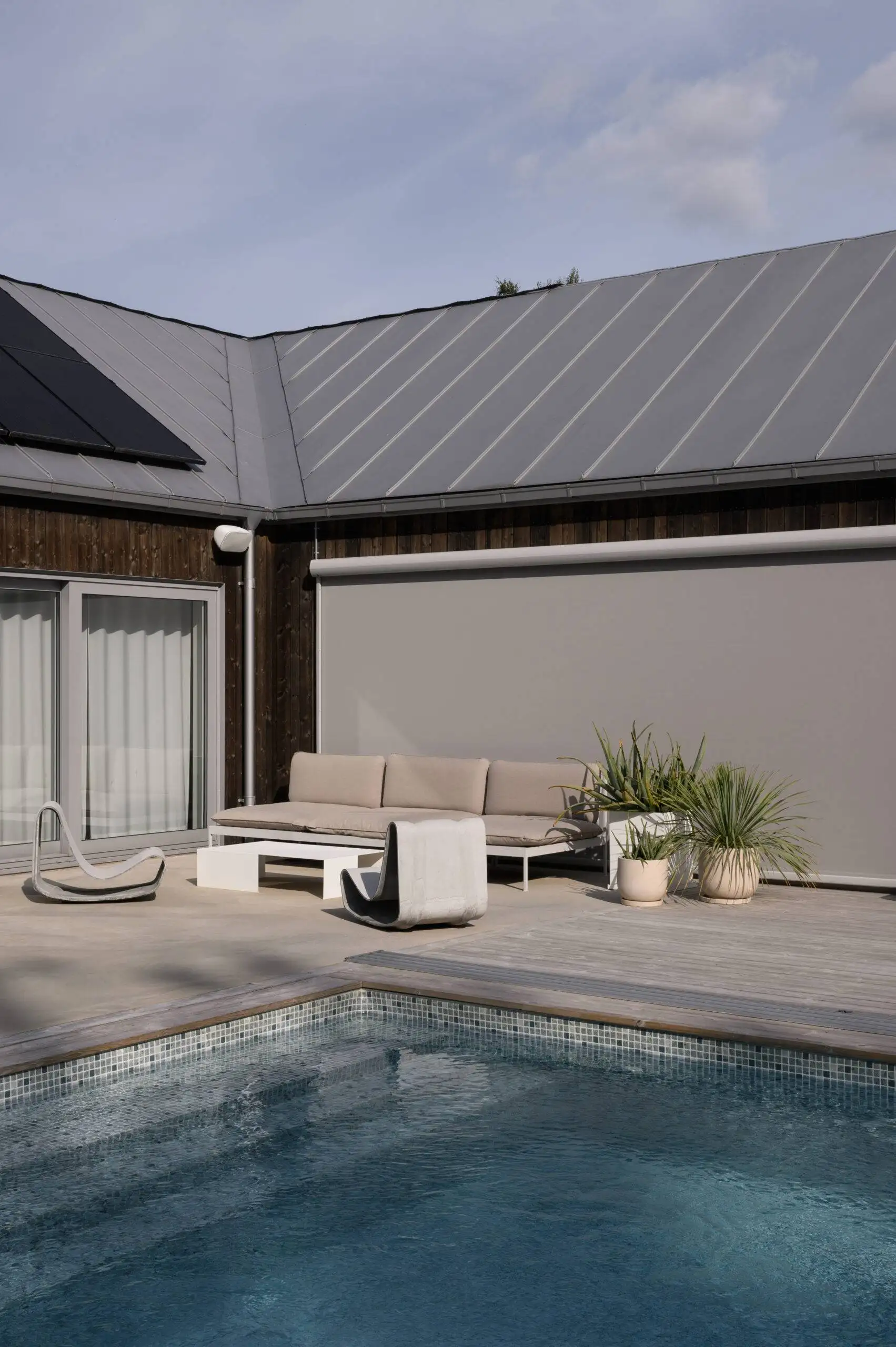 Modern uteplats med pool, beige soffa med småbord och två svängda stolar på träaltan med växter och en nedrullad Styra zipscreen på husets fasad i Stockholm.