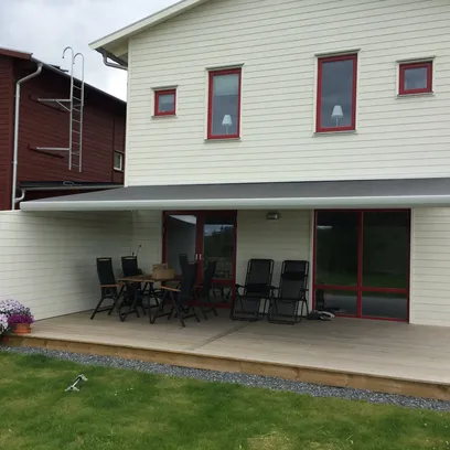 Vitt trähus med rött fönsterfoder, träaltan med utomhusmöbler och en utfälld terrassmarkis ovanför altanen i Stockholm.