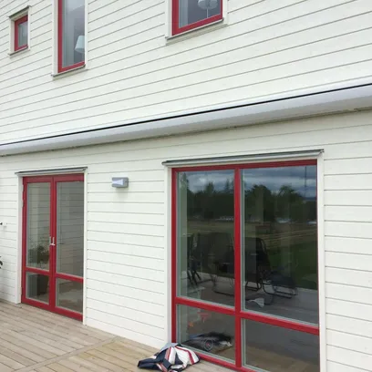 Vit träfasad med röda fönster och glasdörrar mot en trädäckad uteplats med en indragen terrassmarkis i Stockholm.
