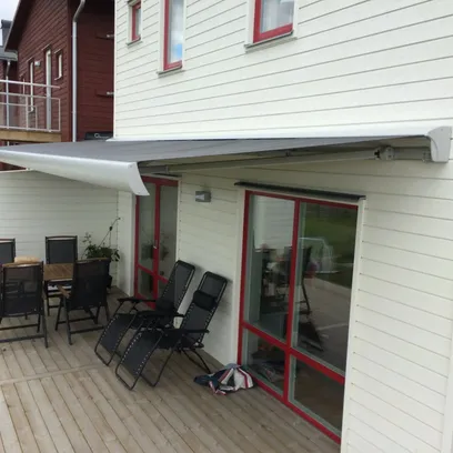Uteplats med träaltan, två svart vilstolar, ett matbord med sex stolar och en utdragbar terrassmarkis på en vit träfasad med röda fönsterkarmar i Stockholm.