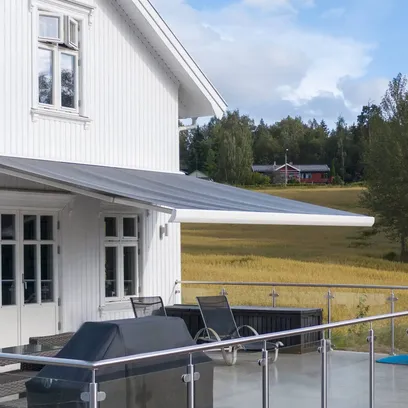 Vit trävilla med utdragen grå terrassmarkis över uteplats med glasräcke, utemöbler och blå barnpool i naturmiljö i Stockholm.