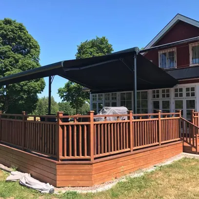 Trädäck med svart terrassmarkis Duo och trästaket vid ett rött och vitt hus i solsken i Stockholm.