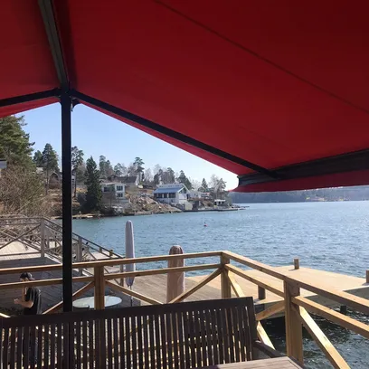Utsikt över en sjö med bryggor, hus vid strandkanten och en röd terrassmarkis Duo ovanför en träbänk på en altan i Stockholm.